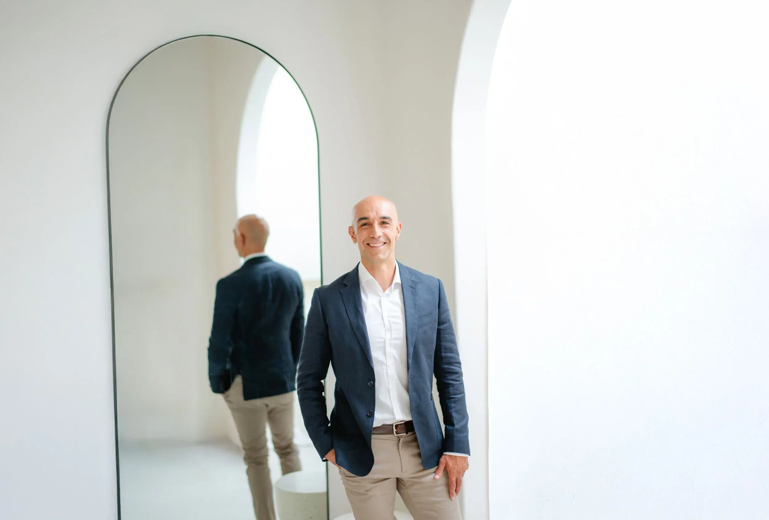Mike McGregor in a navy blazer, beside an arched mirror that shows his reflection.